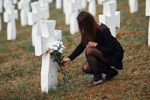 gibt Respekt, indem er Blumen legt. junge Frau in schwarzer Kleidung, die den Friedhof mit vielen weißen Kreuzen besucht. Vorstellung von Begräbnis und Tod foto