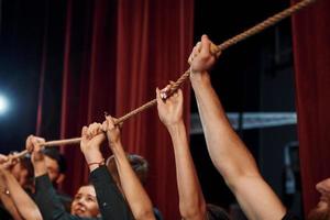 Seil in den Händen über den Köpfen halten. Gruppe von Schauspielern in dunkler Kleidung bei der Probe im Theater foto