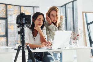 Kamera auf Stativ zeichnet auf. zwei junge freiberuflerinnen, die drinnen im büro mit laptop arbeiten foto