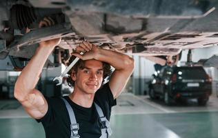 Schöner erwachsener Mann in grauer Uniform, der unter dem Auto steht foto