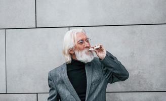 elektronische Zigarette rauchen. Senior Geschäftsmann in formeller Kleidung, mit grauem Haar und Bart ist im Freien foto