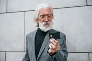 senior geschäftsmann in formeller kleidung, mit grauem haar und bart ist im freien hält kreditkarte foto