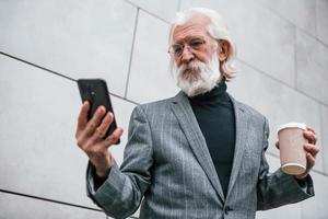 mit Smartphone. Senior Geschäftsmann in formeller Kleidung, mit grauem Haar und Bart ist im Freien foto