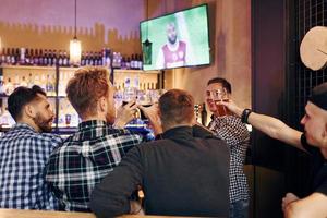 Anfeuern durch Klopfen von Biergläsern. gruppe von menschen zusammen drinnen in der kneipe haben spaß am wochenende foto