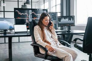 spricht per Telefon. junge börsenmaklerin arbeitet drinnen im büro. viele Displays foto