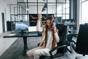 spricht per Telefon. junge börsenmaklerin arbeitet drinnen im büro. viele Displays foto