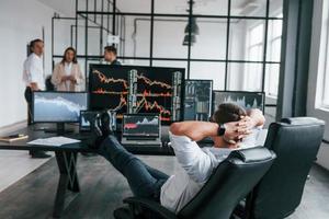 mann sitzt auf stuhl und macht pause. Das Team von Börsenmaklern arbeitet in einem modernen Büro mit vielen Bildschirmen foto