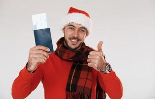 hält Flugticket. Junger gutaussehender Mann in Neujahrskleidung, der drinnen vor weißem Hintergrund steht foto