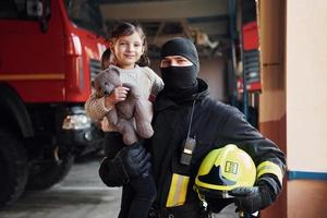 hält Teddybär. glückliches kleines mädchen ist mit männlichem feuerwehrmann in schutzuniform foto