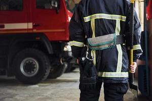 hält axt in den händen. männlicher feuerwehrmann in schutzuniform, der in der nähe von lkw steht foto