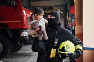 hält Teddybär. glückliches kleines mädchen ist mit männlichem feuerwehrmann in schutzuniform foto