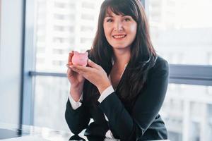 Frau in schwarzer formeller Kleidung ist drinnen im modernen Büro und hält rosa Sparschwein foto