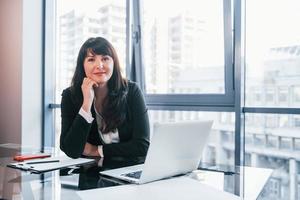 mit Laptop auf dem Tisch. Frau in schwarzer formeller Kleidung ist drinnen im modernen Büro foto