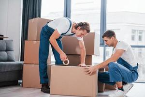 Verpackung der Kiste. Zwei junge Umzugshelfer in blauer Uniform arbeiten drinnen im Raum foto