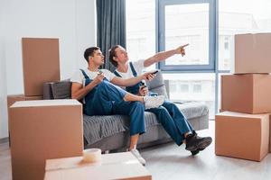 auf dem Sofa sitzen. müde und macht Pause. Zwei junge Umzugshelfer in blauer Uniform arbeiten drinnen im Raum foto