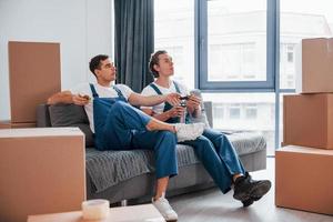 auf dem Sofa sitzen. müde und macht Pause. Zwei junge Umzugshelfer in blauer Uniform arbeiten drinnen im Raum foto