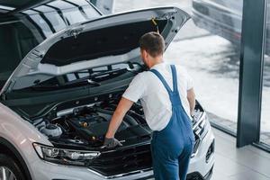 Haube geöffnet ist. junger mann in weißem hemd und blauer uniform repariert autos foto