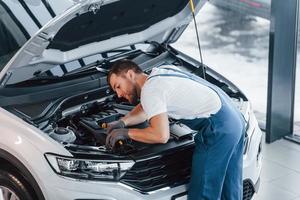 Haube geöffnet ist. junger mann in weißem hemd und blauer uniform repariert autos foto