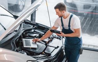 nutzt Laptop. junger mann in weißem hemd und blauer uniform repariert autos foto