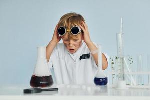 verängstigter kleiner Junge im Mantel, der mit Geräten einen Wissenschaftler im Labor spielt foto