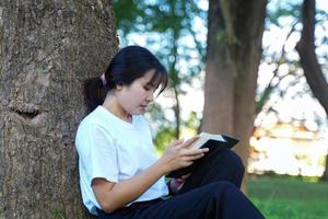 asiatische Frau, die mit dem Rücken gegen einen Baum sitzt und ein Buch liest. Konzept. asiatische frau, die outdoor-aktivitäten macht, wie bücher lesen, arbeiten, ein picknick mit der familie machen. weicher und selektiver fokus foto