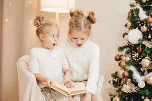 2 blonde Mädchen sitzen auf einem Sessel am Weihnachtsbaum und lesen ein Buch. gemütliche Abende mit einem Buch am Baum. Überraschungsgefühl bei Kindern foto