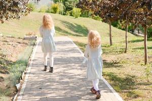 2 kleine blonde Mädchen mit lockigem Haar gehen in einem Sommerpark spazieren. süße und kleine Mädchen auf der Straße. 2 Schwestern, die in passenden Kleidern und weißen Strumpfhosen nach draußen gehen foto