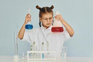 Kleines Mädchen im Mantel, das einen Wissenschaftler im Labor spielt, indem es Ausrüstung verwendet foto