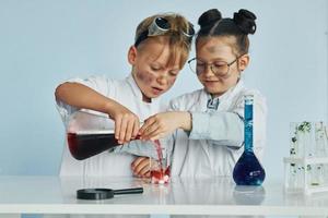 Kleines Mädchen und Junge in weißen Mänteln spielen einen Wissenschaftler im Labor, indem sie Geräte verwenden foto