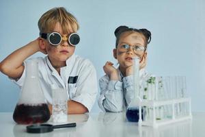 Mädchen mit Jungen, die zusammenarbeiten. Kinder in weißen Mänteln spielen einen Wissenschaftler im Labor, indem sie Geräte verwenden foto