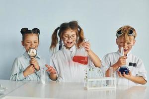 glückliche Freunde lächeln. Kinder in weißen Mänteln spielen einen Wissenschaftler im Labor, indem sie Geräte verwenden foto