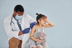 Junger afroamerikanischer Arzt, der einem kleinen Mädchen im Krankenhaus eine Injektion gibt foto