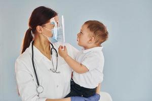 junge krankenschwester im weißen kittel und mit stethoskop hält kleinen jungen in den händen foto