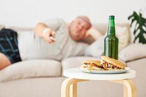 Hamburger und Bier. Mann liegt auf dem Sofa. lustiger übergewichtiger mann in lässiger kleidung ist drinnen zu hause foto