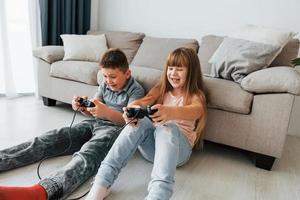 auf dem Boden sitzen und Videospiel spielen. Kinder, die sich tagsüber gemeinsam im Wohnzimmer amüsieren foto