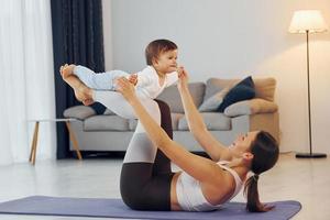 frau, die sich auf matte legt und kleines mädchen hält. Mutter mit ihrer kleinen Tochter ist zusammen zu Hause foto