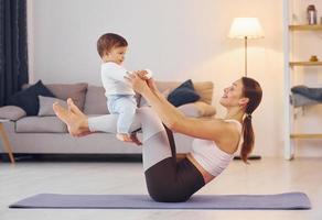 Seitenansicht einer Frau, die Übungen mit einem kleinen Mädchen macht. Mutter mit ihrer kleinen Tochter ist zusammen zu Hause foto