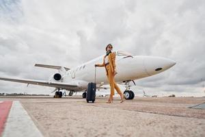 elegante Kleidung. junge frau mit gepäck ist draußen in der nähe des flugzeugs foto