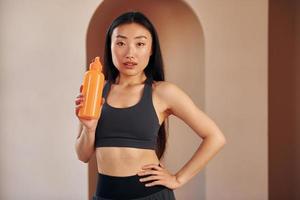 mit orangefarbener Flasche. junge ernsthafte asiatische frau, die drinnen steht foto