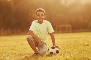 sitzt mit Fußball. afroamerikanisches kind hat tagsüber im sommer spaß auf dem feld foto