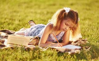 Kleines Mädchen liest Buch, während es sich im Freien auf das Gras des Feldes legt foto