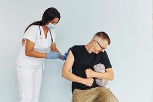 Mann hält Teddybär während des Verfahrens. Arzt in Uniform, der dem Patienten eine Impfung vornimmt foto