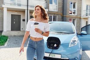 Auto aufladen. Junge Frau in Freizeitkleidung mit ihrem Elektromobil tagsüber im Freien foto