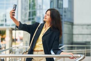 Telefon in der Hand halten. Frau in formeller Kleidung, die tagsüber draußen in der Stadt steht foto