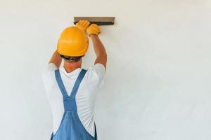 Stehen in der Nähe der weißen Wand. junger mann, der tagsüber in uniform auf dem bau arbeitet foto