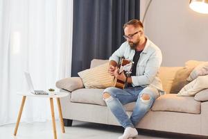 gemütliche Atmosphäre. Mann in Freizeitkleidung und mit Akustikgitarre ist drinnen foto