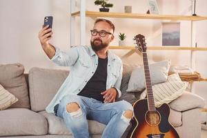 auf dem Sofa sitzen. Mann in Freizeitkleidung und mit Akustikgitarre ist drinnen foto