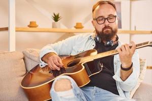 kreative beschäftigung. Mann in Freizeitkleidung und mit Akustikgitarre ist drinnen foto