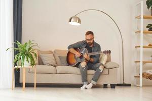 Üben von Songwriting-Fähigkeiten. Mann in Freizeitkleidung und mit Akustikgitarre ist drinnen foto