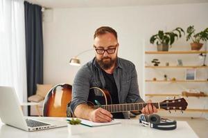 einige Ideen schreiben. Mann in Freizeitkleidung und mit Akustikgitarre ist drinnen foto